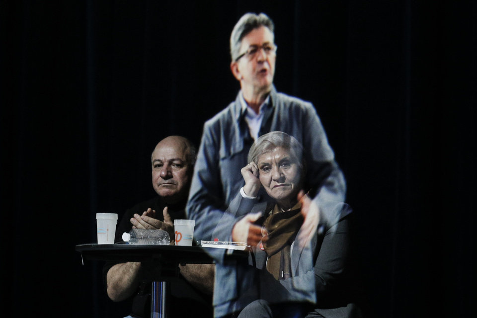 People sit at a table on stage and look towards the hologram of hard-left French presidential candidate Jean-Luc Melenchon, as he speaks to supporters who are gathered in Saint-Denis, near Paris, Sunday, Feb. 5, 2017. As Melenchon holds a rally in Lyon Sunday, a hologram of him is being projected by satellite to crowds in Paris. (AP Photo/Kamil Zihnioglu)